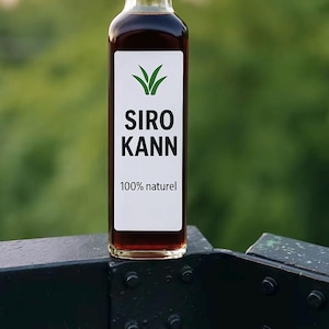 May include: A clear glass bottle filled with a dark liquid, featuring a white label with the text "SIRO KANN" and "100% naturel". The bottle has a black cap and is set against a blurred green background.