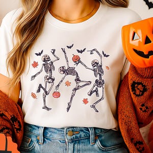 May include: A white t-shirt with a graphic of dancing skeletons, bats, and autumn leaves. The image includes a pumpkin-shaped mug and a tote bag with a similar design. The theme is Halloween.