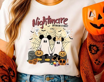 Może przedstawiać: Biała koszulka z grafiką o tematyce Halloween. Projekt przedstawia dwie postacie duchów z uszami Myszki Miki i Minnie, pajęczynę, dynie i napis "Nightmare on Main Street". Na zdjęciu znajduje się pomarańczowy kubek w kształcie dyni.