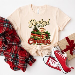 A beige t-shirt with a graphic design of stacked books and a mug, along with a red plaid shirt, a gift box, and a pair of red sneakers.