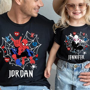 May include: Black t-shirts with Spider-Man and Spider-Gwen designs. The Spider-Man shirt has the name "JORDAN" in white, while the Spider-Gwen shirt has "JENNIFER". Both shirts feature a web design with hearts and spiders.