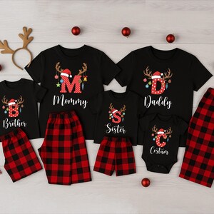 May include: Matching family Christmas pajamas sets in black with red and black plaid bottoms. Each shirt has a letter and the word "Mommy", "Daddy", "Brother", "Sister", and "Costum". The shirts have a reindeer and Santa hat design.