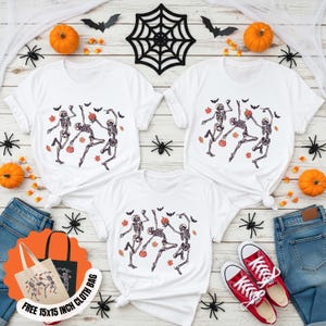 May include: White t-shirts featuring a Halloween skeleton dance design. The image includes pumpkins, bats, spiders, and a tote bag that says "FREE 15x15 INCH CLOTH BAG". Red trainers and blue jeans are also in the image.
