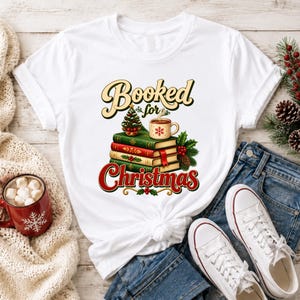 A white t-shirt with a graphic design of a stack of books and a mug, along with Christmas-themed items such as pine cones and a mug of hot chocolate.