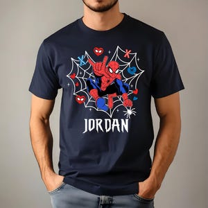 May include: Dark blue t-shirt with a Spider-Man graphic set within a white web. The superhero is depicted in a dynamic pose, with the name "JORDAN" in white script below. The design includes red, blue, and white accents.