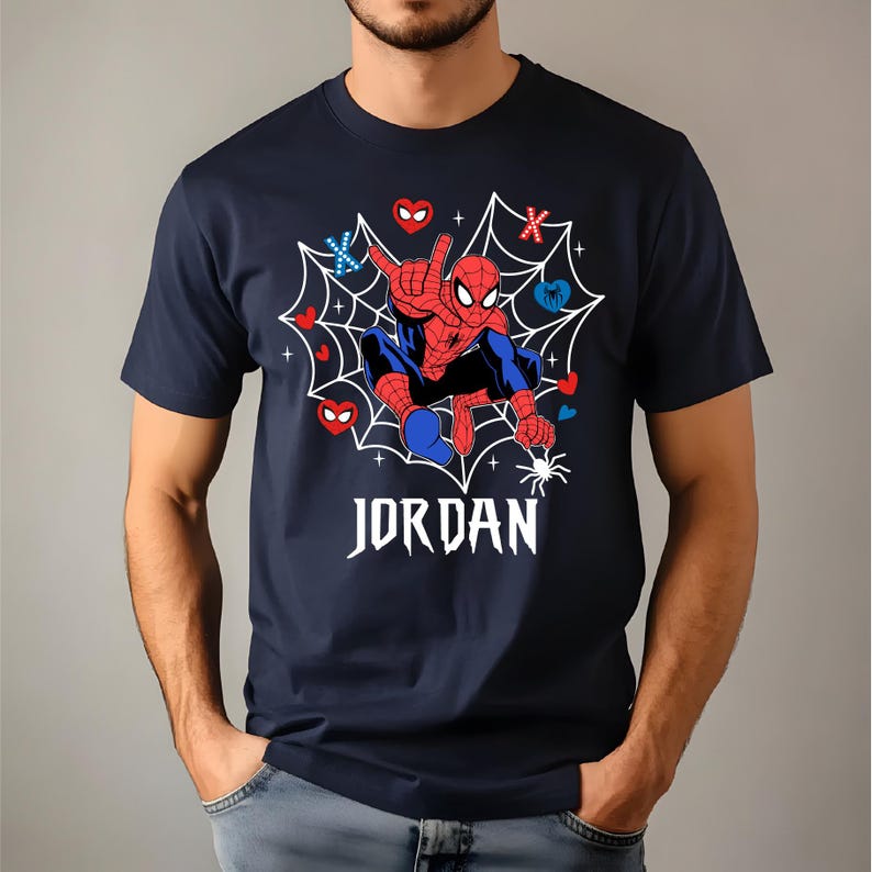 May include: Dark blue t-shirt with a Spider-Man graphic set within a white web. The superhero is depicted in a dynamic pose, with the name "JORDAN" in white script below. The design includes red, blue, and white accents.