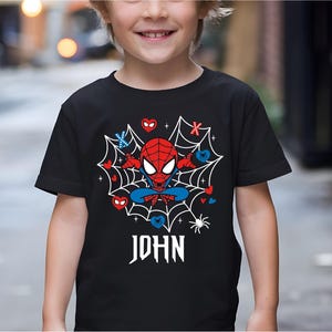 May include: Black t-shirt with a Spider-Man cartoon design. The graphic shows a red and blue Spider-Man character in a web with hearts and the name "JOHN" in white gothic lettering. The shirt has short sleeves.