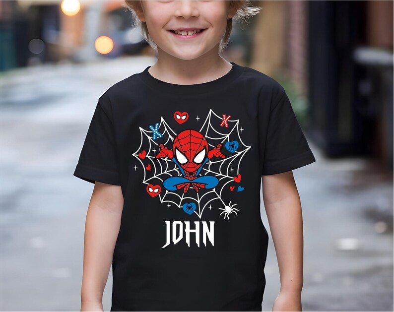 May include: Black t-shirt with a Spider-Man cartoon design. The graphic shows a red and blue Spider-Man character in a web with hearts and the name "JOHN" in white gothic lettering. The shirt has short sleeves.