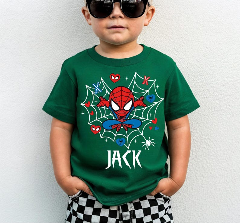 May include: A green t-shirt with a Spider-Man design. The graphic shows a red and blue Spider-Man character in a web, with hearts, snowflakes, and the name "JACK" in white. The child is wearing black sunglasses.