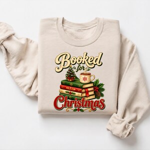 A beige sweater with a graphic design on it, featuring a stack of books, a mug, and a Christmas tree.