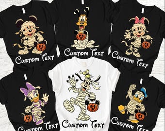 May include: A collection of six short-sleeve t-shirts in black and white, each featuring a different Disney character wrapped in mummy bandages and holding a pumpkin bucket. The text "CUSTOM TEXT" is printed in a script font below each character.