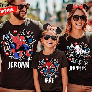 May include: Black t-shirts with Spider-Man and Spider-Gwen designs, personalised with names in a white gothic font. The shirts have a spiderweb graphic with hearts and spider accents. The family is wearing matching Mickey Mouse ears.