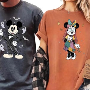 Könnte beinhalten: Ein dunkelgraues T-Shirt mit einer Grafik von Mickey Mouse im Nadelstreifenanzug mit Geistern und Fledermäusen sowie ein orangefarbenes T-Shirt mit einer Grafik von Minnie Mouse im Patchwork-Kleid mit Herbstblättern und Sternen.