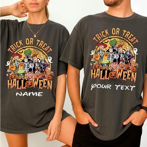 Pode incluir: T-shirts cinzentas escuras com um gráfico temático de Halloween. O design apresenta personagens de desenhos animados, abóboras e o texto "Trick or Treat" e "Happy Halloween". Uma t-shirt tem "NAME" e a outra "your TEXT".