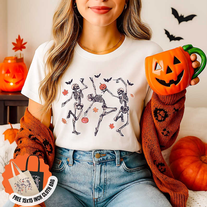 May include: A white t-shirt with a graphic of dancing skeletons, bats, and autumn leaves. The image includes a pumpkin-shaped mug and a tote bag with a similar design. The theme is Halloween.