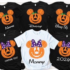 May include: Five short-sleeved t-shirts, four black and one white, each featuring a pumpkin graphic shaped like a mouse head. The black shirts display orange pumpkins with mouse faces and text reading Name, Daddy, Disney Trip, and Costume Text. The white shirt features an orange pumpkin with a purple polka-dot bow and the text Mommy and 2026.
