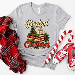 A gray t-shirt with a Christmas-themed graphic design, including a stack of books, a mug, and a Christmas tree, along with a pair of red and white striped candy canes and a red and white plaid shirt.