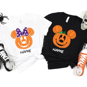 May include: Two Halloween-themed t-shirts, one white and one black, each featuring a smiling pumpkin design with Mickey Mouse ears. The white shirt has a purple polka-dot bow. The word "NAME" is printed below the pumpkin. Black and orange sneakers are visible.