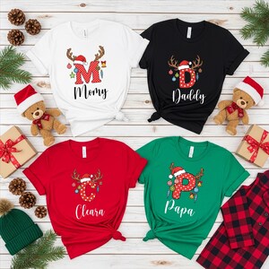 May include: Four Christmas-themed t-shirts in white, black, red, and green. Each shirt features a letter and name, with reindeer antlers and festive decorations. The shirts are tied at the waist and surrounded by holiday decor.