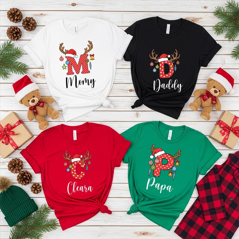 May include: Four Christmas-themed t-shirts in white, black, red, and green. Each shirt features a letter and name, with reindeer antlers and festive decorations. The shirts are tied at the waist and surrounded by holiday decor.