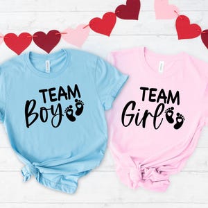 May include: Two short-sleeved t-shirts, one light blue and one pink, with the words "TEAM Boy" and "TEAM Girl" printed in black script. Each shirt features a pair of baby footprints. A garland of red and pink hearts hangs in the background.