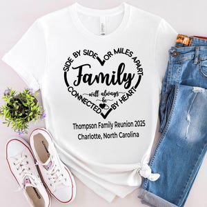 May include: A white short-sleeve t-shirt featuring a black heart-shaped graphic with the text Side by Side or Miles Apart Family will always be connected by heart, Thompson Family Reunion 2025, and Charlotte, North Carolina printed on the front.