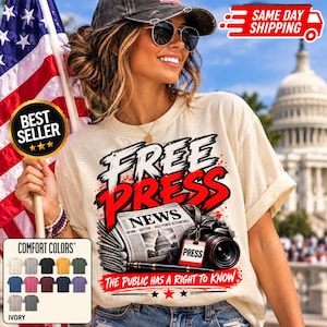 May include: An ivory t-shirt featuring a graphic print with the text "FREE PRESS" in large black and red letters. Below, a newspaper illustration shows the text "NEWS" and "THE PUBLIC HAS A RIGHT TO KNOW" alongside a camera and a pen.