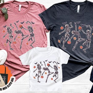 May include: Three t-shirts in various colours, including a dusty rose, charcoal, and white, each with a dancing skeleton design with pumpkins and bats. A free 38x38 cm cloth bag with the same design is also included.