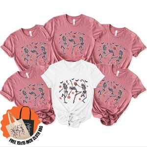 May include: Six t-shirts, five in a dusty rose hue and one white, are displayed. Each shirt has a dancing skeleton design with pumpkins, bats, and leaves, ideal for Halloween. A free 38x38 cm cloth bag is included.