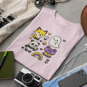 May include: A light pink t-shirt featuring a cartoon fox, panda, ghost, and frog with hearts and a rainbow. Other items include a brown hat, sunglasses, a camera, a notebook, and a tote bag.