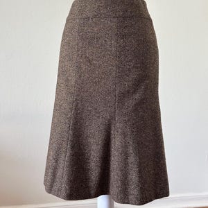 May include: A brown tweed skirt with a classic A-line silhouette. The skirt features a subtle textured pattern and a tailored fit, suitable for various occasions. The skirt is displayed on a white mannequin.