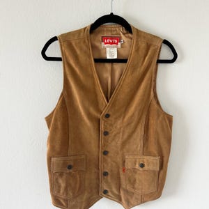 Può includere: Un gilet in pelle scamosciata marrone chiaro con scollo a V, due tasche frontali e chiusura con bottoni. Il gilet ha un'etichetta Levi's ed è appeso a una gruccia nera contro un muro bianco. Il gilet è uno stile classico.