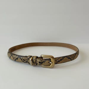 May include: A slim belt featuring a tan and black snakeskin pattern throughout the strap. The belt is secured with a matte gold-toned metal buckle and a matching metal keeper loop, providing a functional closure for this accessory item.