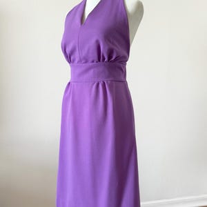 May include: A solid purple sleeveless dress featuring a halter neckline, a wide fitted waistband with subtle gathering above and below the band, and a knee-length skirt. The fabric has a textured surface and the dress is displayed on a mannequin.