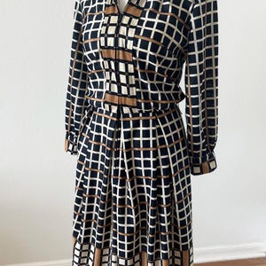 May include: A long-sleeved midi dress featuring a black, white, and tan grid pattern. The dress has a collared neckline, a button-down front placket, and a pleated skirt. The fabric displays a consistent geometric check design throughout the garment.