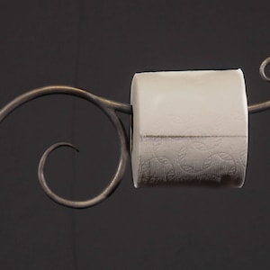 May include: A metal toilet paper holder with a decorative scroll design. The holder is made of dark metal and has a single roll of toilet paper on it.