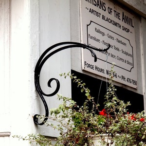 May include: A black metal sign with the text "Artisans of the Anvil, Artist Blacksmith Shop" and a list of services offered. The sign is mounted on a white wall with a black metal plant hanger attached to the wall. A green plant with red flowers hangs from the plant hanger.