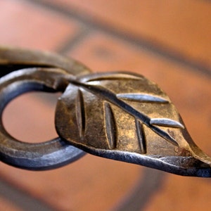 May include: Close-up of a hand-forged metal leaf and ring. The leaf has detailed veining and is attached to a curved stem. The metal has a dark, aged finish, suggesting a rustic or vintage style.