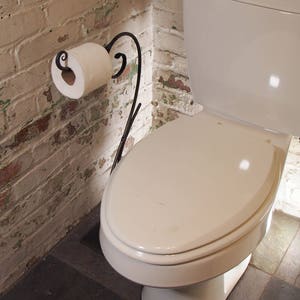 May include: A white toilet with a white toilet seat in a bathroom with a white brick wall and a black metal toilet paper holder with a roll of toilet paper.