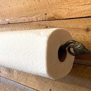 May include: A white paper towel roll mounted on a wooden surface. The roll is held in place by a decorative, dark metal leaf-shaped holder. The paper has a textured pattern.