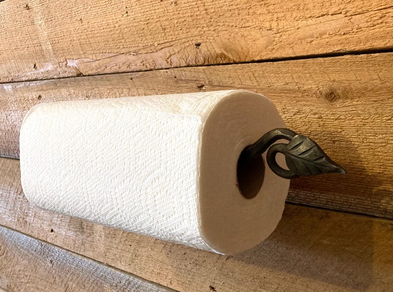 May include: A white paper towel roll mounted on a wooden surface. The roll is held in place by a decorative, dark metal leaf-shaped holder. The paper has a textured pattern.