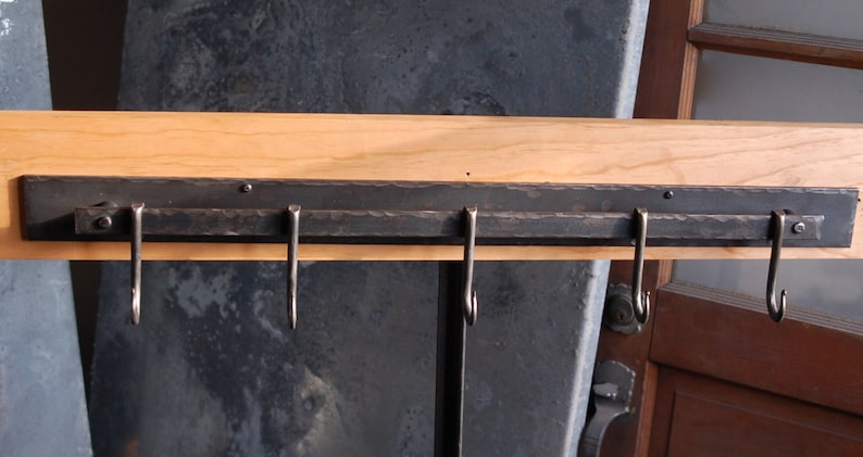 Op de afbeelding: Een zwarte metalen rek met vijf haken gemonteerd op een houten oppervlak. Het rek is ontworpen om keukengerei of andere items aan op te hangen.