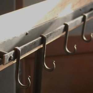 Op de afbeelding: Een metalen rek met vier haken bevestigd aan een houten oppervlak. De haken zijn gemaakt van zwart metaal en zijn aan het einde gebogen. Het rek is aan de muur bevestigd.