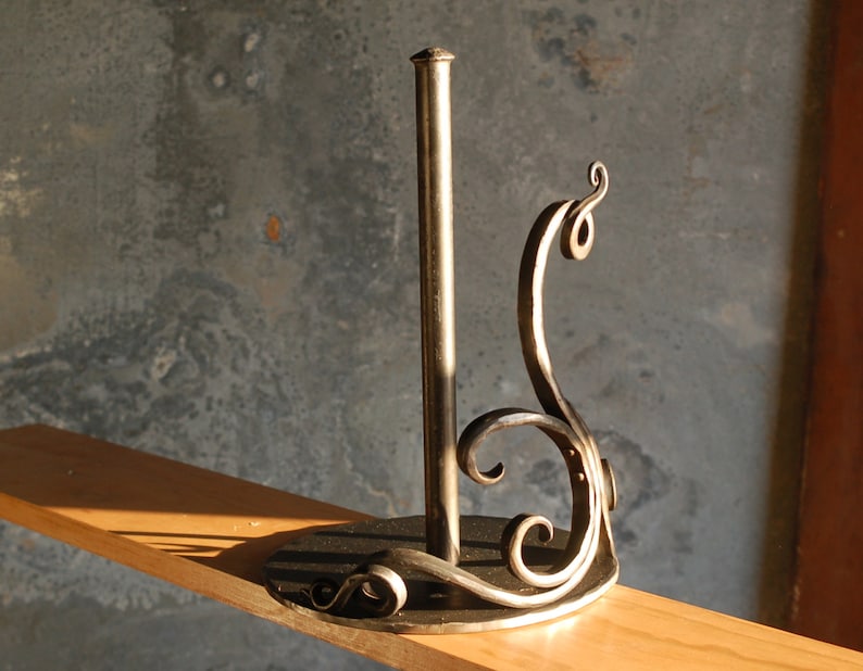 May include: A metal paper towel holder with a decorative scroll design. The holder is made of dark metal and has a round base.