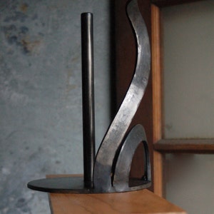 May include: A black metal paper towel holder with a curved design. The holder is mounted on a wooden shelf.