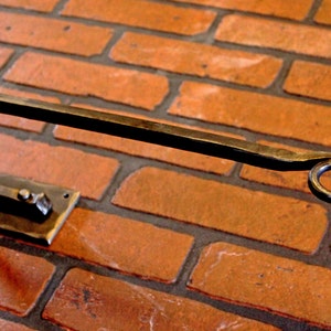 May include: Hand-forged metal towel rack mounted on a brick wall. The rack features a rectangular base with two rivets, a curved arm, and a leaf-shaped detail at the end. The metal has a dark, rustic finish.