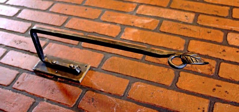May include: Hand-forged metal towel rack mounted on a brick wall. The rack features a rectangular base with two rivets, a curved arm, and a leaf-shaped detail at the end. The metal has a dark, rustic finish.