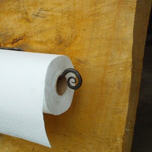 May include: A white paper towel roll mounted on a black metal paper towel holder with a decorative scroll design. The holder is attached to a wooden surface.