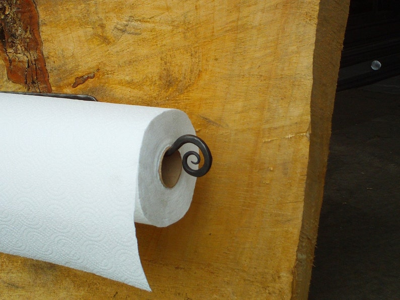 May include: A white paper towel roll mounted on a black metal paper towel holder with a decorative scroll design. The holder is attached to a wooden surface.