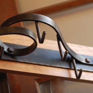 May include: A metal coat rack with a decorative, curved design. The rack has multiple hooks for hanging coats, hats, or other items. The rack is mounted on a wooden surface.